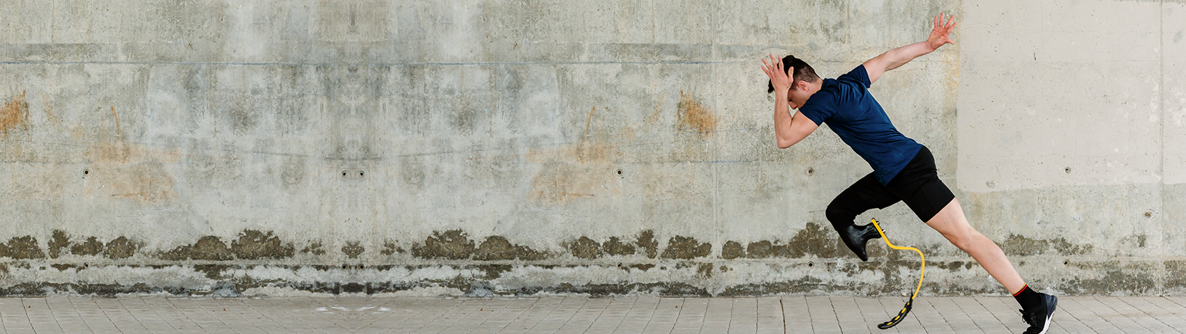 Full body side view of strong determined male athlete with leg prosthesis ready for sprint on pavement against concrete wall